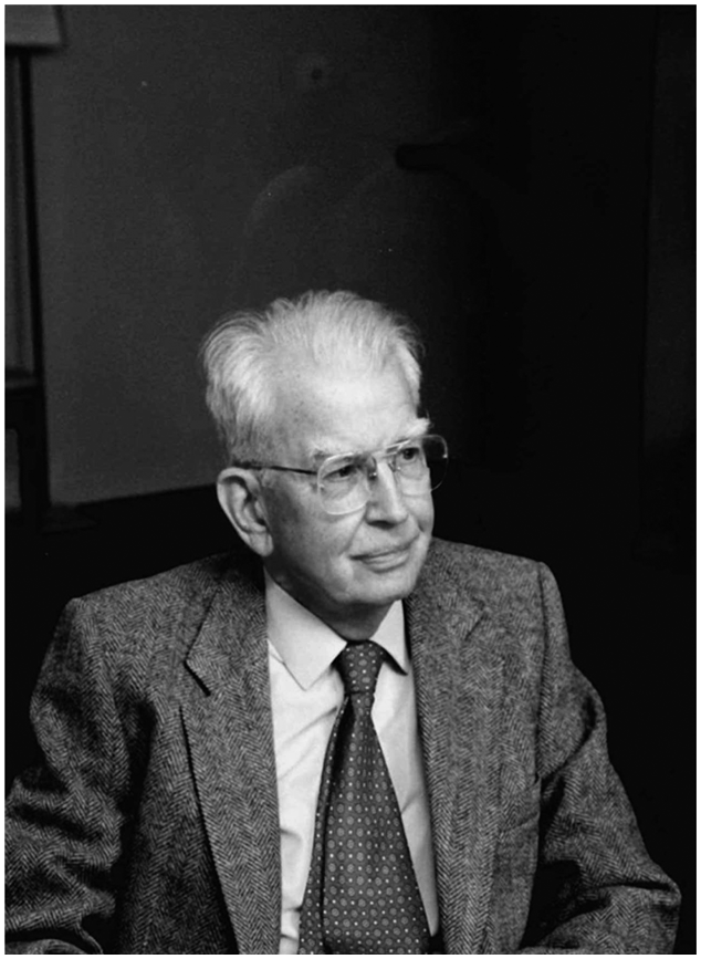 Close-up portrait of an older man with glasses and white hair. He is wearing a suit jacket and tie, and looking away from the camera. The background is dark and out of focus.