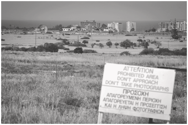 A photograph of a landscape with a warning sign in the foreground and buildings in the background. The signboard reads Attention, Prohibited Area Don't Take Photographs, in English as well as in Greek.