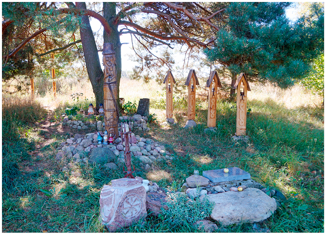 A photograph of a shady grove with brightly lit fields behind it. A central sculpture is carved in wood, showing a figure holding a drinking horn. Candles are in front of it, and carved stones hold offerings of fruits and coins.
