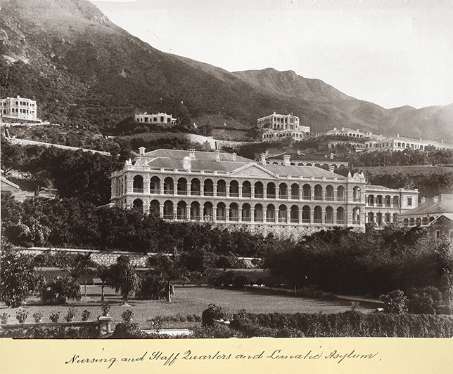The photo shows the asylum, which is built onto a hillside. The asylum has an arched verandah façade. Behind the asylum are smaller buildings with a similar style. In front of the asylum, there is a park lined with bushes, trees, and plants.