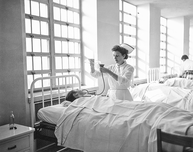 A nurse administers insulin to a patient. The patient is lying flat in a bed. Their face cannot be seen. The nurse pours a clear bottle of the medication into a funnel. The funnel is attached to a tube and connected to the patient.