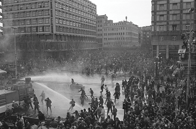 A scene depicts the brutal, massive protests in Belgrade on 9 March 1991, where hundreds of thousands of people took to the streets to protest Slobodan Milošević’s rule and the imminent start of the civil wars. See long description.