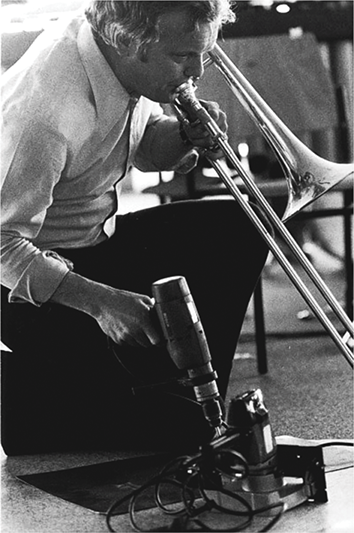 Vinko Globokar performing at the Music Biennale Zagreb in May 1977. He is wearing a shirt and pants, kneeling and playing a trombone with his left hand while simultaneously using a power drill with his right.