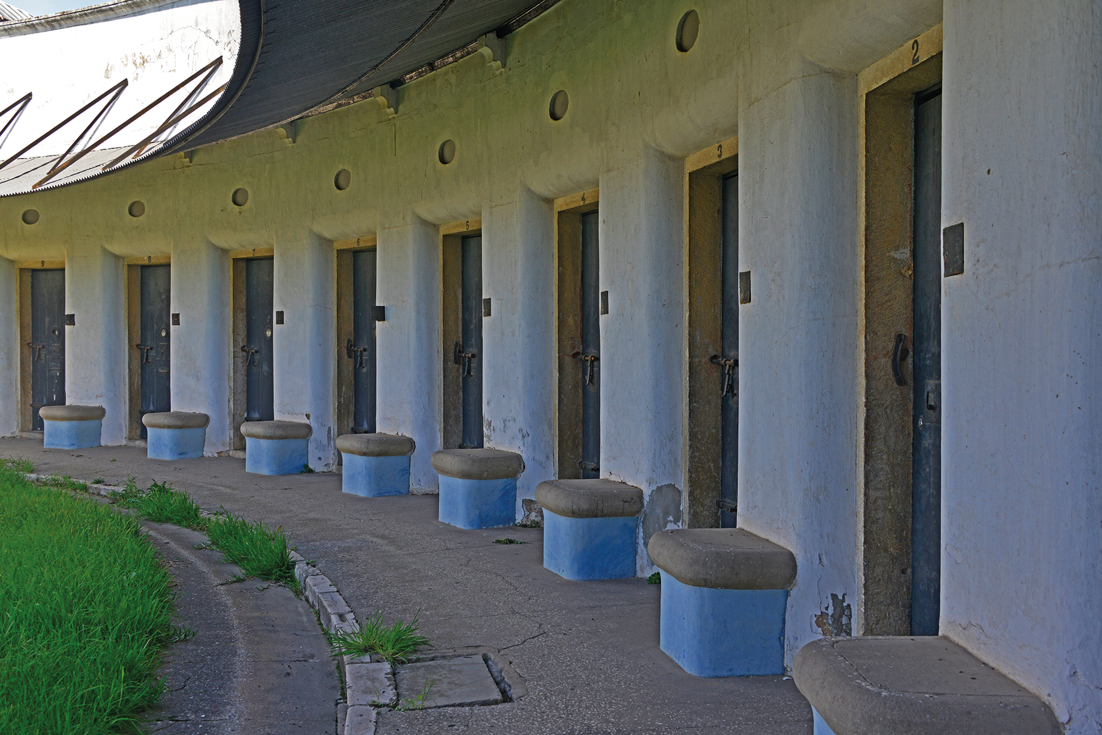 A row of doors with benches in front of each, set against a white wall with a curved roof overhead.