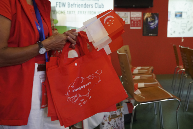 A woman, dressed in red and white, holding several red and white Singapore flags, alongside red tote bags with ‘Singapore’ written on them.