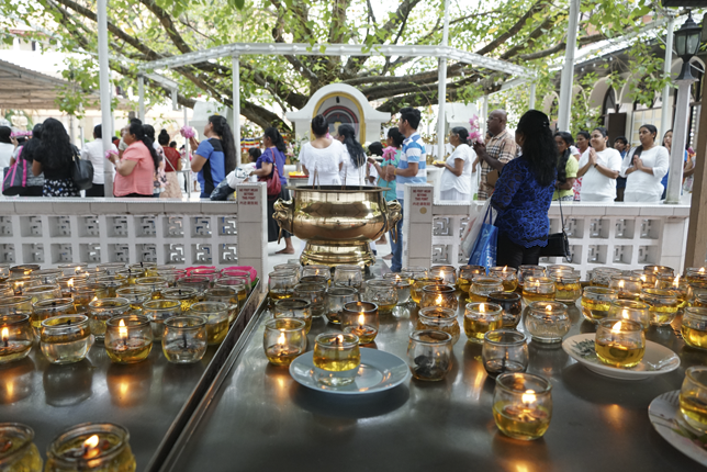 Several lit candles are in the foreground. A woman looks on at other people in prayer, walking under and around a large Bodhi tree.