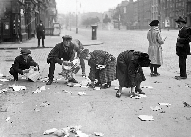 A group of four people in the foreground picking up scraps from a Dublin street. A man and a woman are talking in the right rear. Another man stands in the left background.