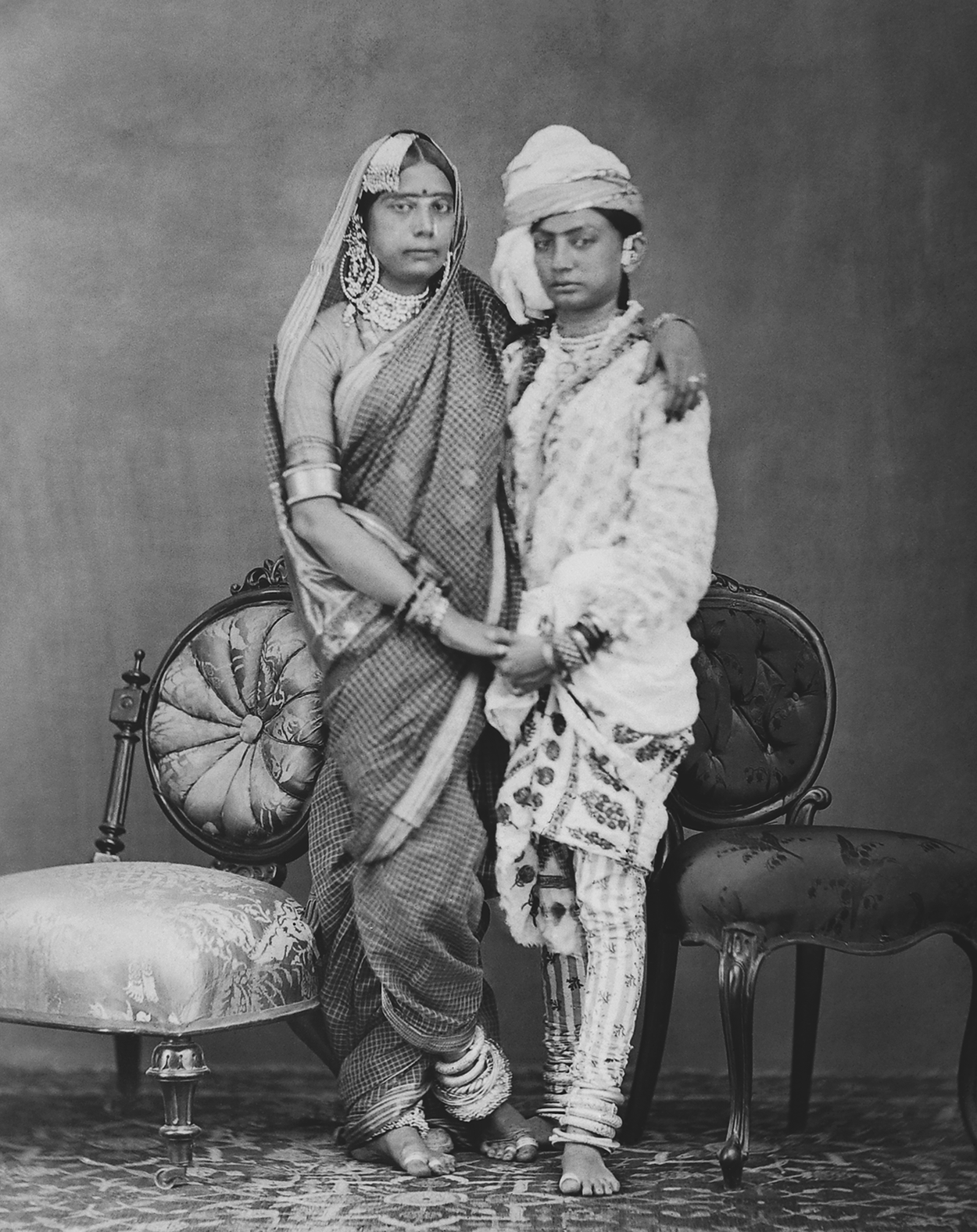 Nanhi Jan and Munna Jan posing together in traditional attire, standing between two chairs. See long description.