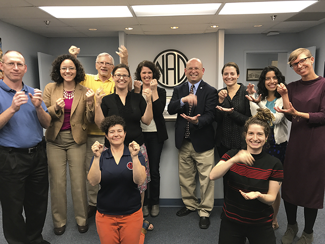 Eleven people standing or kneeling in the NAD headquarters with the NAD logo on the back wall. They are all looking at the camera and smiling while signing their preference for sign or bank in A S L.