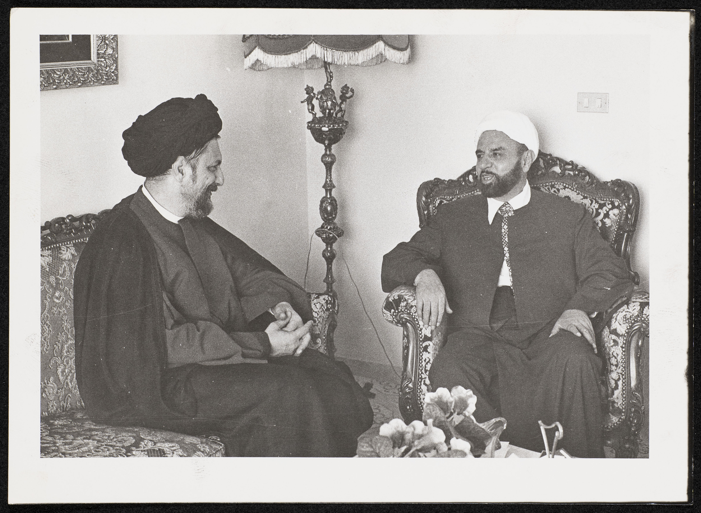 Two men in traditional attire sitting and conversing in a decorated room.
