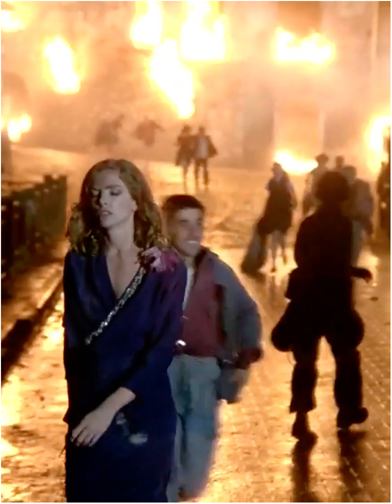 A cinematic scene with a woman and a child walking on a street with fires in the background, surrounded by several people running away from the flames. See long description.