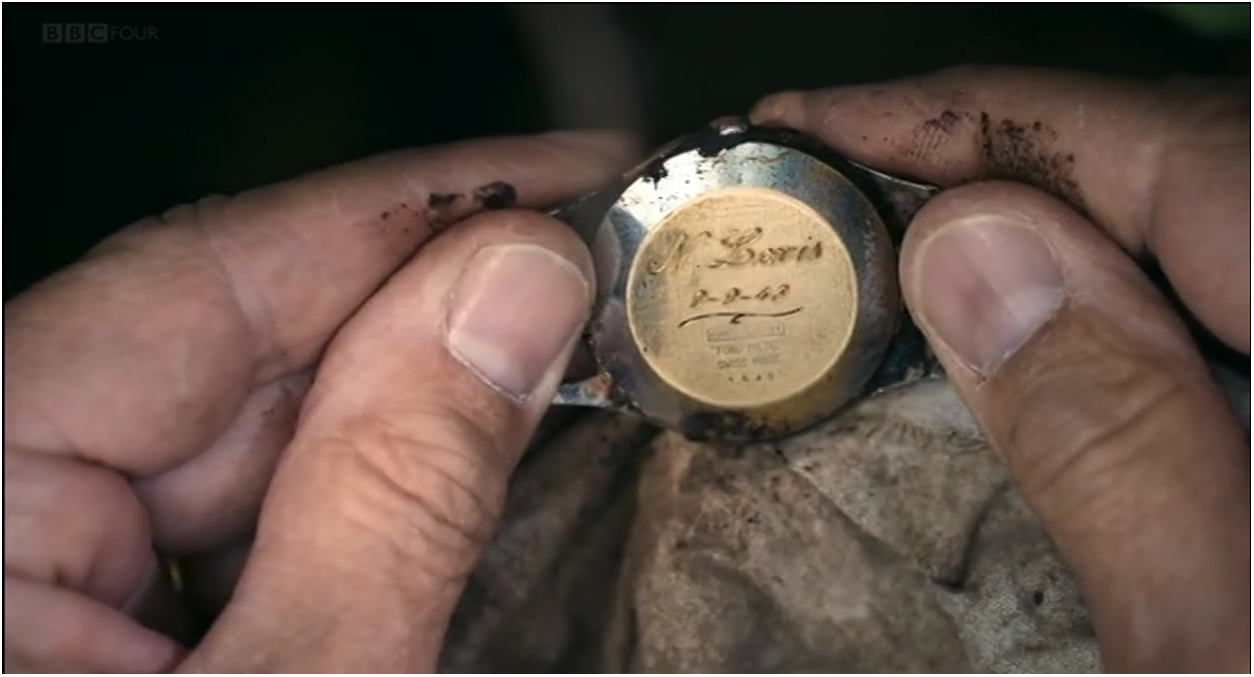 Two hands holding a watch with engraved text on the back, partially covered by a cloth. See long description.