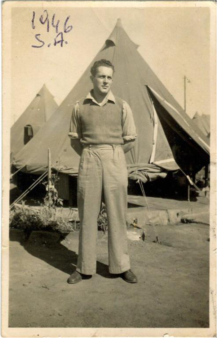 A man standing in front of tents with '1946 S.A.' written on the photo. He is wearing a sweater over a collared shirt and trousers, with hands behind his back. See long description.