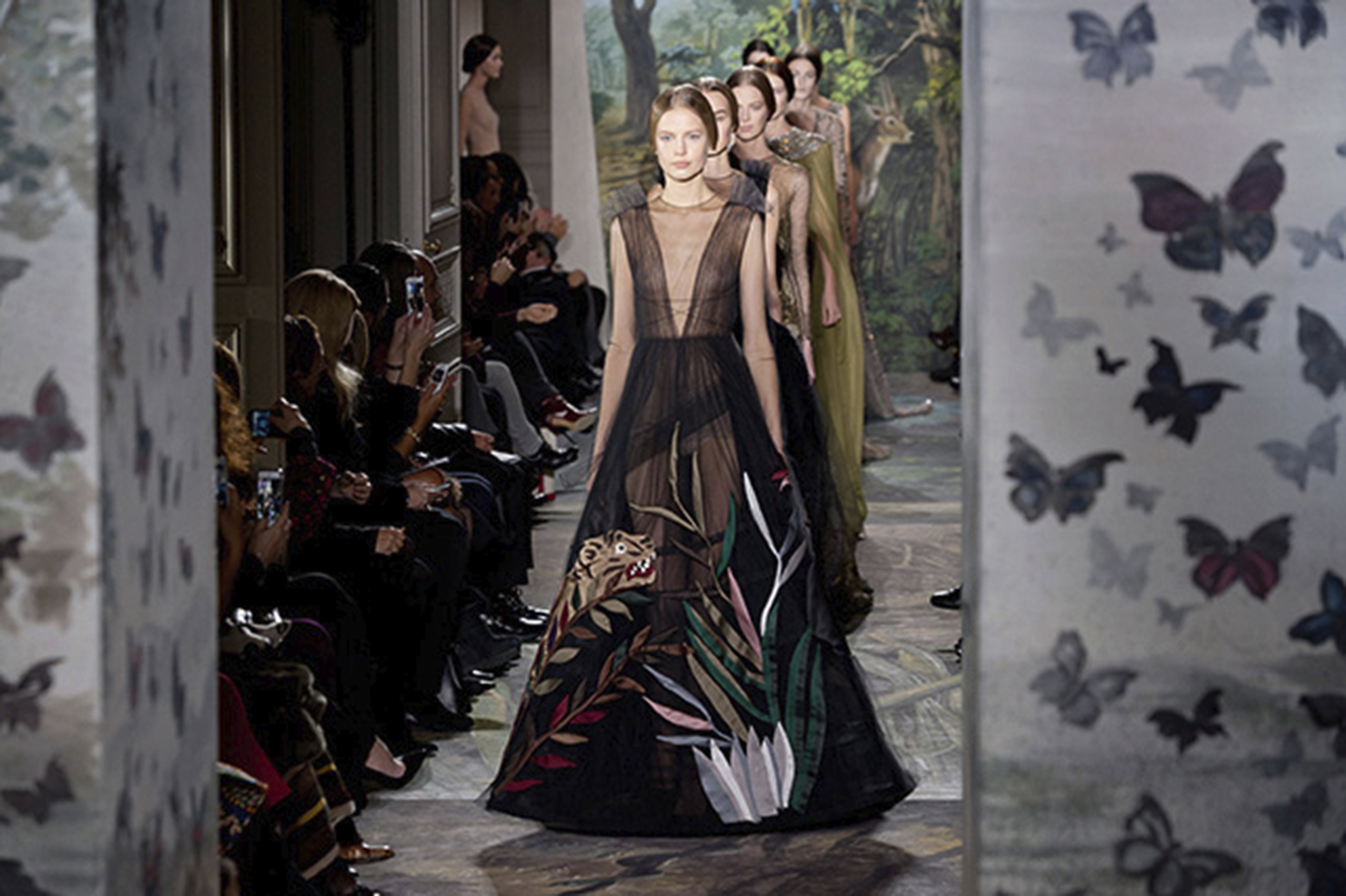 Photo of a fashion runway with models in nature-themed couture gowns, audience seated on both sides, and butterfly motifs on foreground panels. See long description.