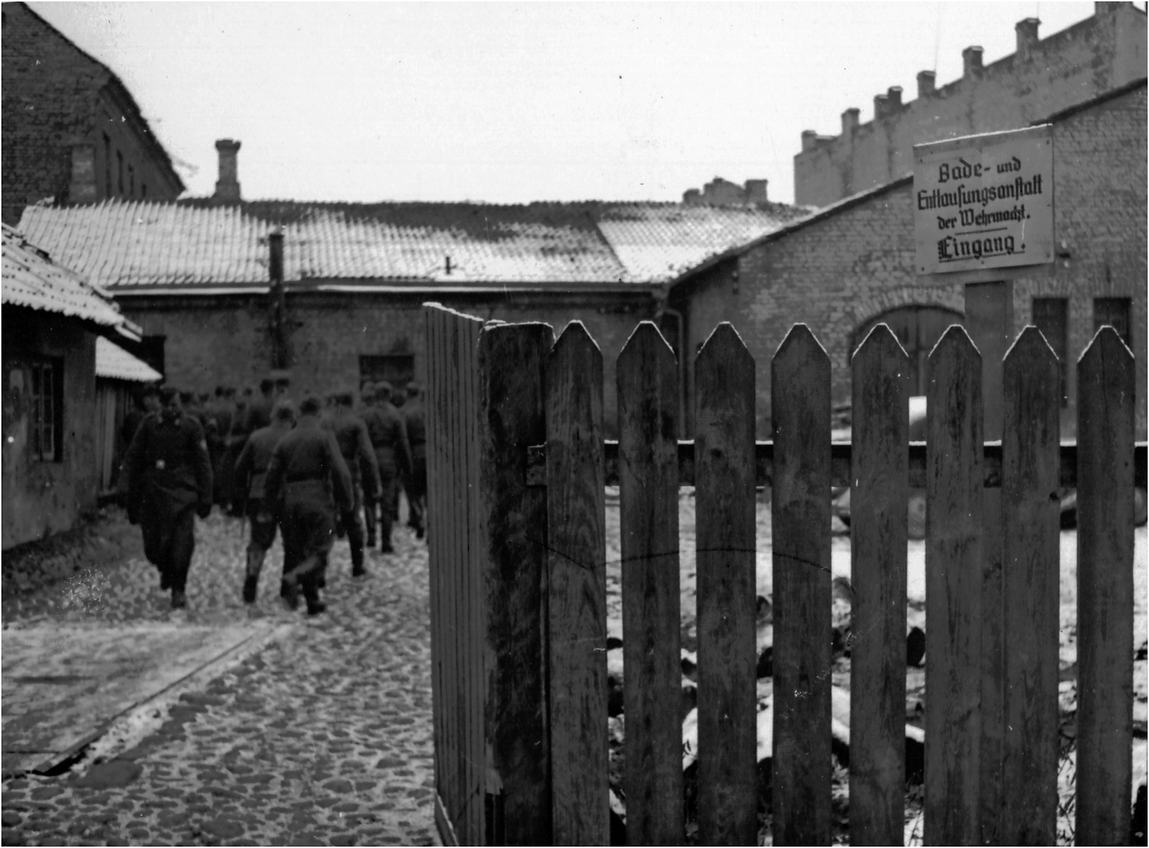Black and white photo of soldiers walking toward a building. A wooden fence and a German sign reading Bade- und Entlausungsanstalt der Wehrmacht. Eingang. are in the foreground. Snow dusts the ground and rooftops.