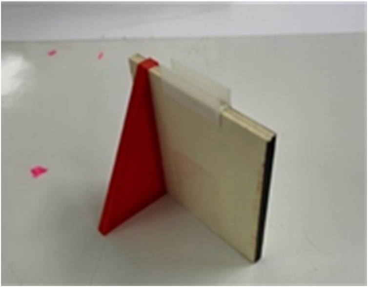 A photo of a vertical wooden board held upright by a red triangular plastic brace on the left side. A small translucent clip is attached to the top edge of the board.
