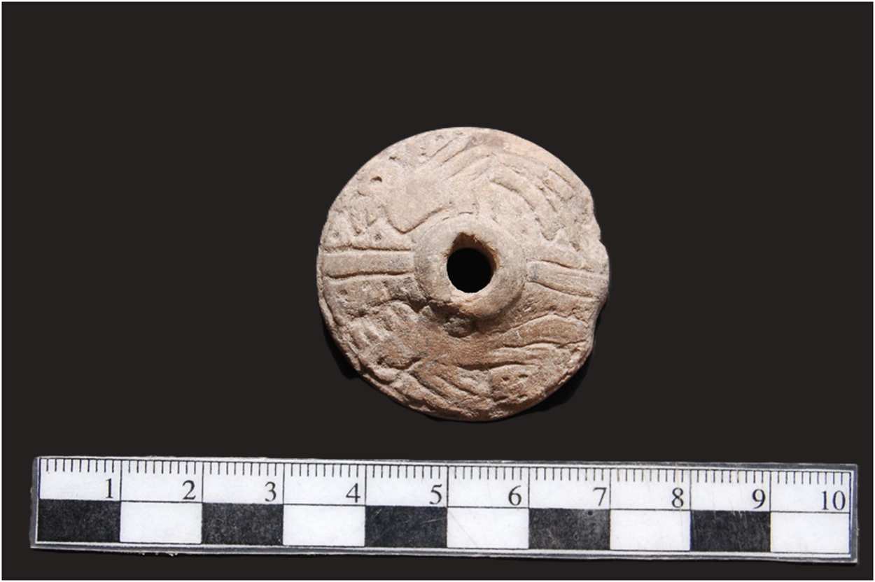 A spindle whorl with intricate carvings next to a ruler showing measurements. See long description.