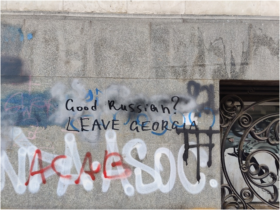Photo of a stone wall with black spray-painted text reading Good Russian? LEAVE GEORGIA. Below this, larger white and red graffiti tags are partially visible, including the letters N A C A S O C.