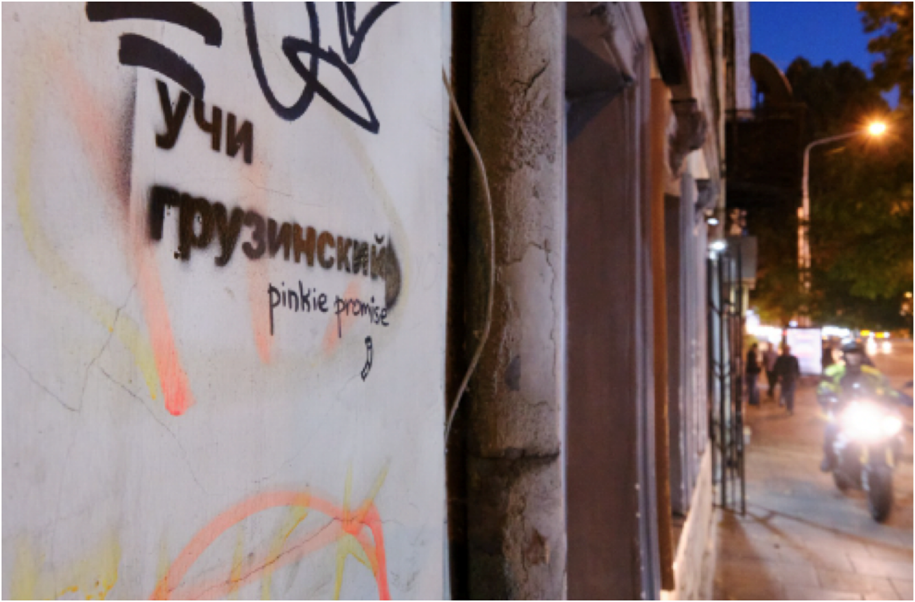 A photo of a white wall with black stenciled Russian text and the English phrase pinkie promise. In the background, a street at dusk features a motorcyclist with a bright headlight and pedestrians on a sidewalk.