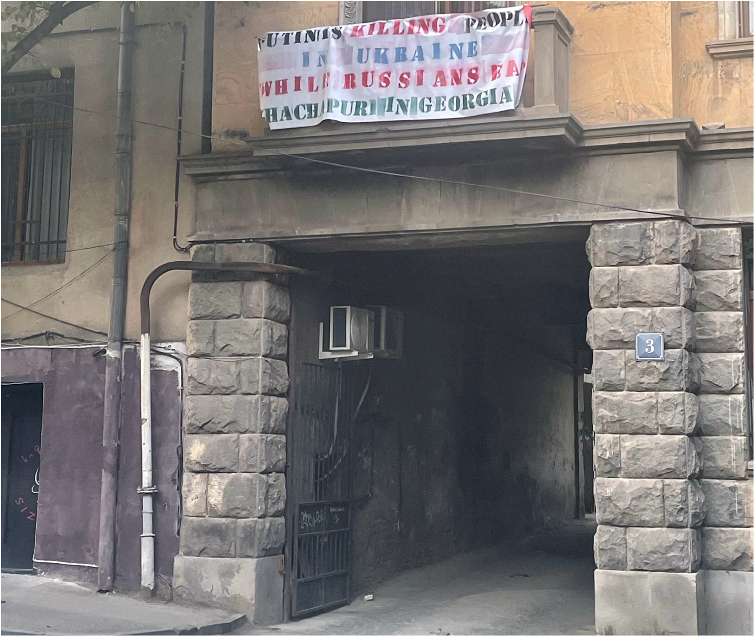 Photo of a white banner hanging from a stone balcony above an arched driveway. The banner text reads Putin is killing people in Ukraine while Russians eat Khachapuri in Georgia in red and blue capital letters.