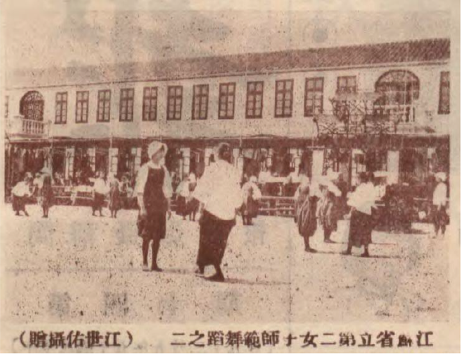 Photo showing female students in uniform skirts and blouses dancing in formation outdoors, with a two-story building with many windows in the background.