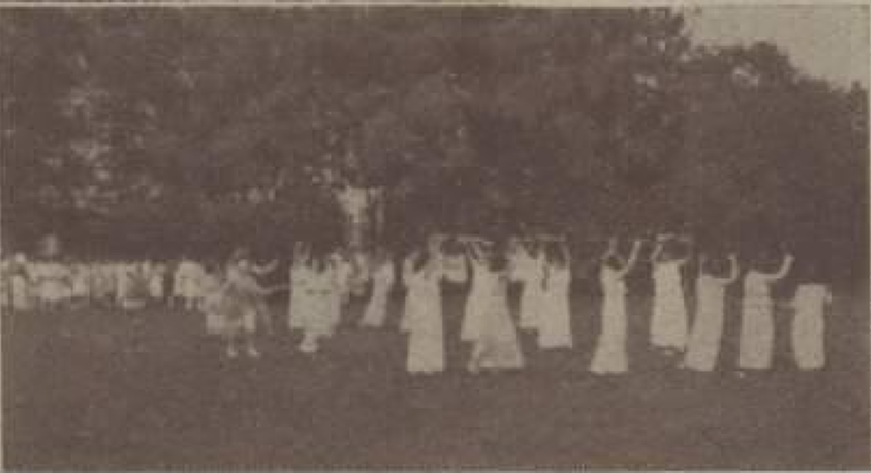 Photo showing a group of girls in white dresses dancing outdoors in rows with arms raised, with trees in the background.