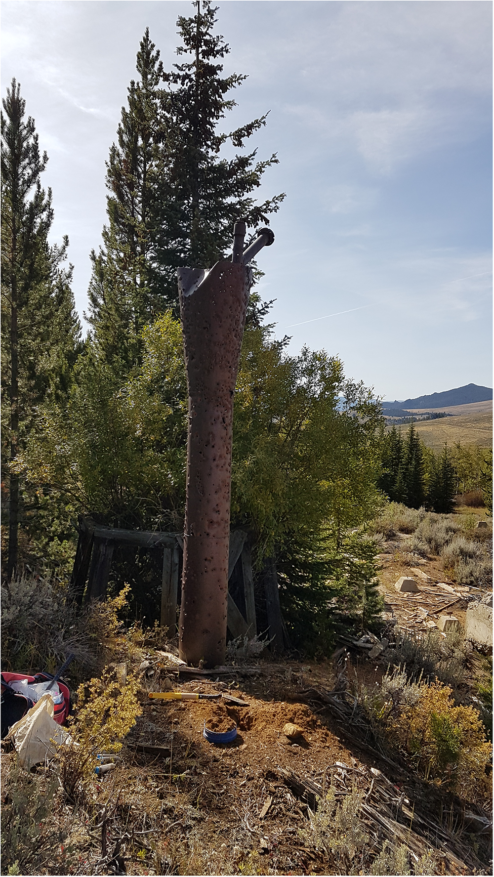 A tall, rusted metal structure stands among tailing piles and trees in a rural landscape. See long description.