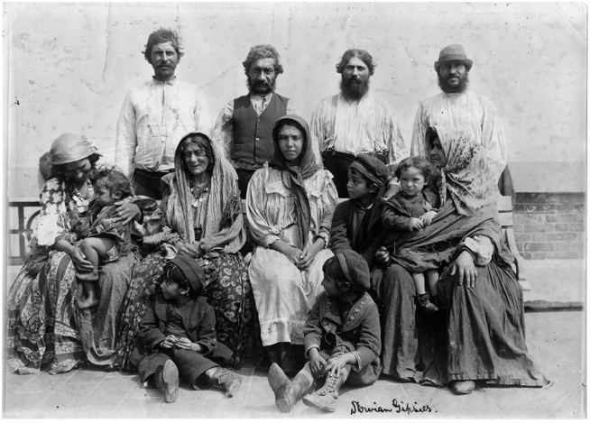 A photo of a large group of Romani people, including men, women, and children, pose outdoors. Handwritten text at the bottom right of the photo reads, “Servian Gipsies”.