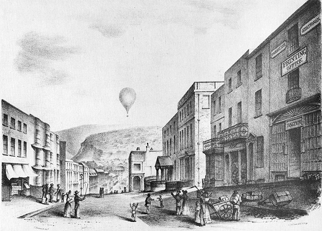 A nineteenth-century street scene at the top of a hill, depicting people walking up and down a high street. A hot air balloon floats in the sky, with hills visible in the background.