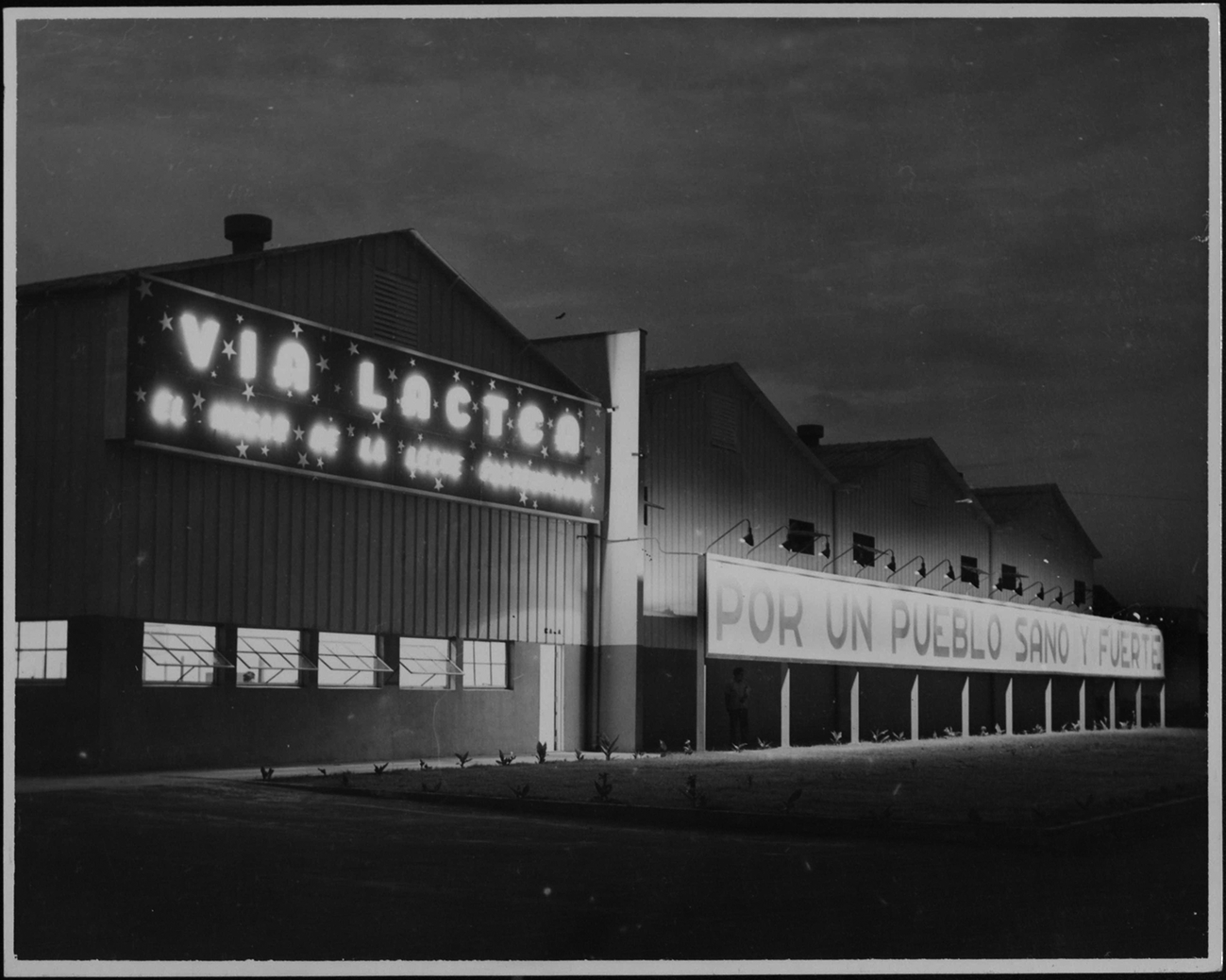 Black and white photo of the Via Lactea factory at night. A glowing neon sign reads VIA LACTEA above a row of windows. To the right, a long banner under spotlights reads POR UN PUEBLO SANO Y FUERTE.