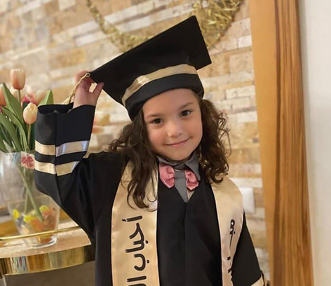 A five-year-old girl standing, wearing a graduation cap and gown with gold trim and a gold scarf around her shoulders, hand on cap, smiling.