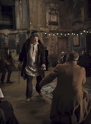 An elderly actor in a simple costume portrays King Lear in a semi-derelict chapel space. The Gothic architecture of the former asylum is visible behind the performer.