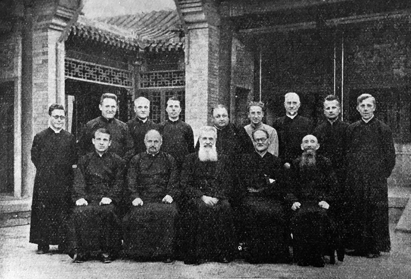 Black and white photo of Father Jozef Vandeputte seated at the center of a group of thirteen men in clerical robes. They are arranged in two rows before a traditional Chinese building with ornate lattice windows and a tiled roof in Peiping.