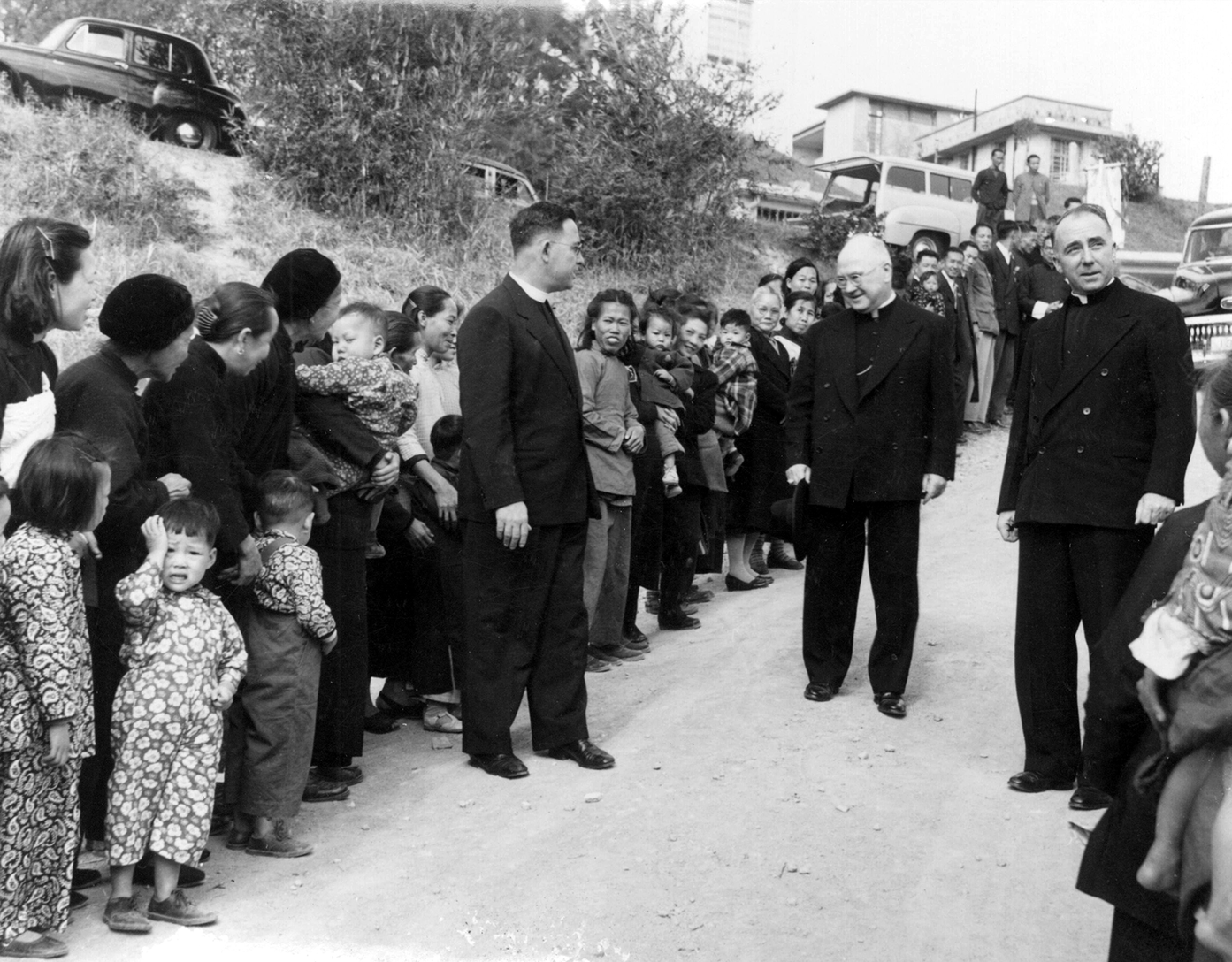 Black and white photo of Msgr. Martin T. Gilligan and Cardinal Francis Spellman standing on a dirt road in a refugee camp. They face a large group of women and children lined up along a grassy hillside with cars parked above.