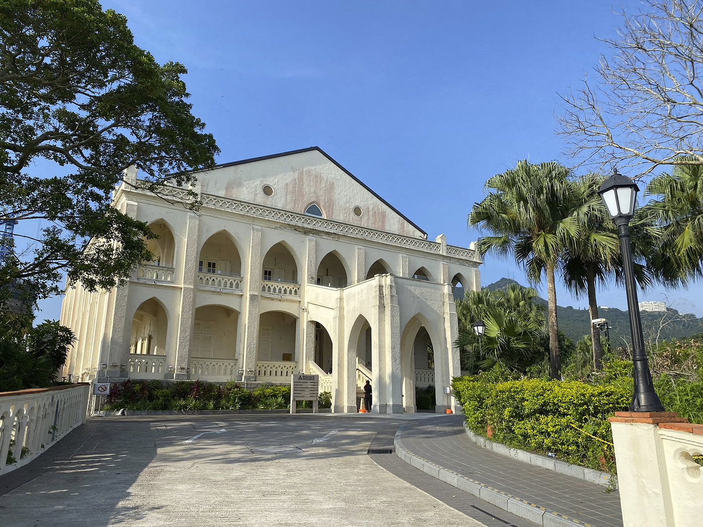 A photo of a two-story cream-colored building with Neo-Gothic pointed arches and a gabled roof. A paved driveway leads to the entrance, flanked by lush green trees and palm trees under a clear blue sky.