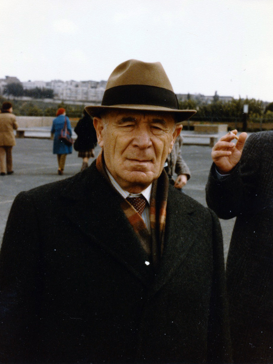 An elderly man wearing a hat and coat stands outdoors in a public area.