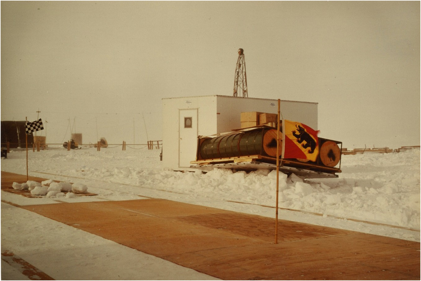 A snowy landscape with a small building, equipment and a flag.