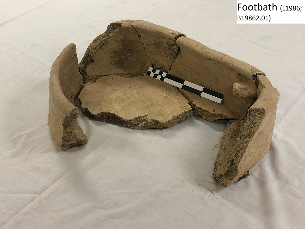 A broken clay footbath with a scale for measurement.