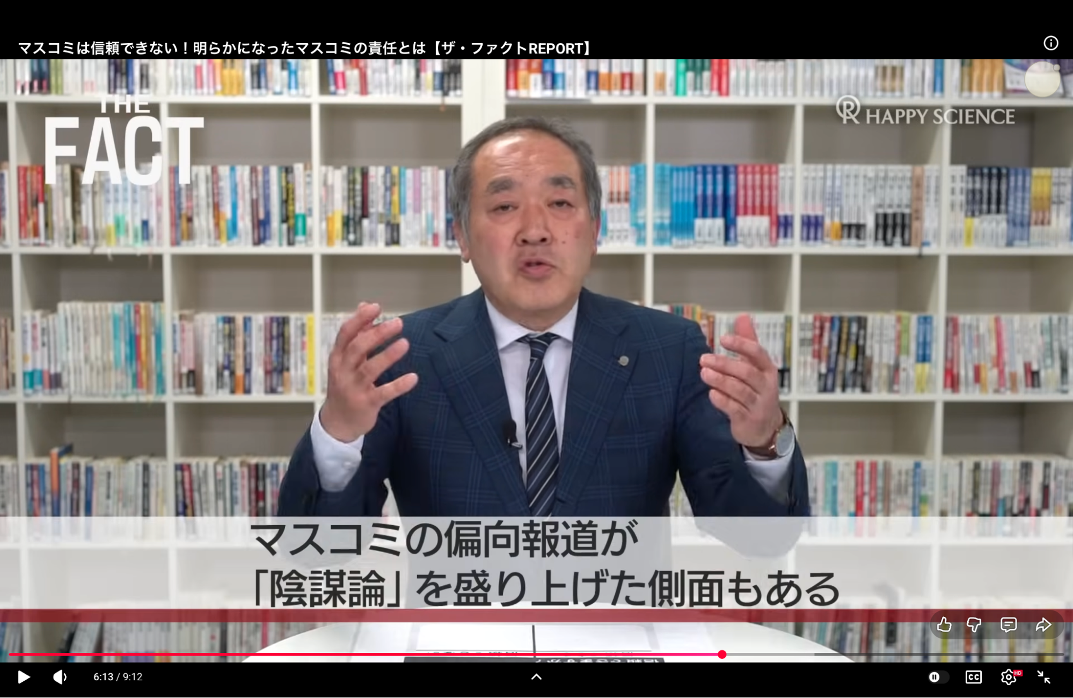A man in a suit and tie gestures while speaking in front of a bookshelf.