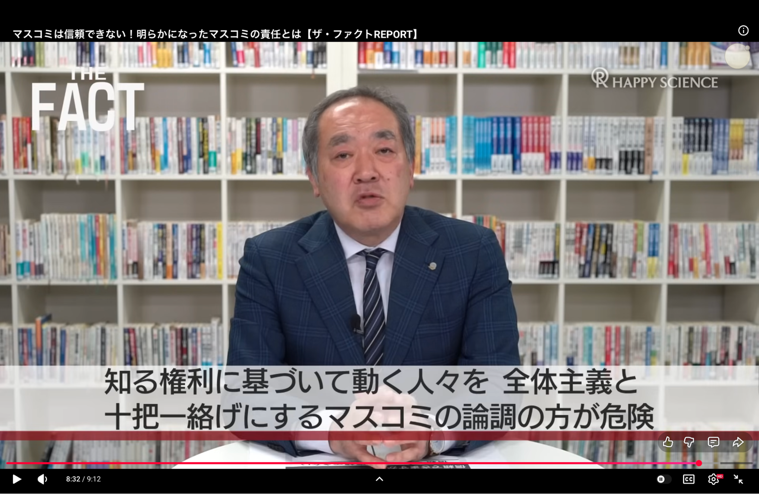 A man in a suit and tie sits in front of a bookshelf, speaking critically about the mass media.