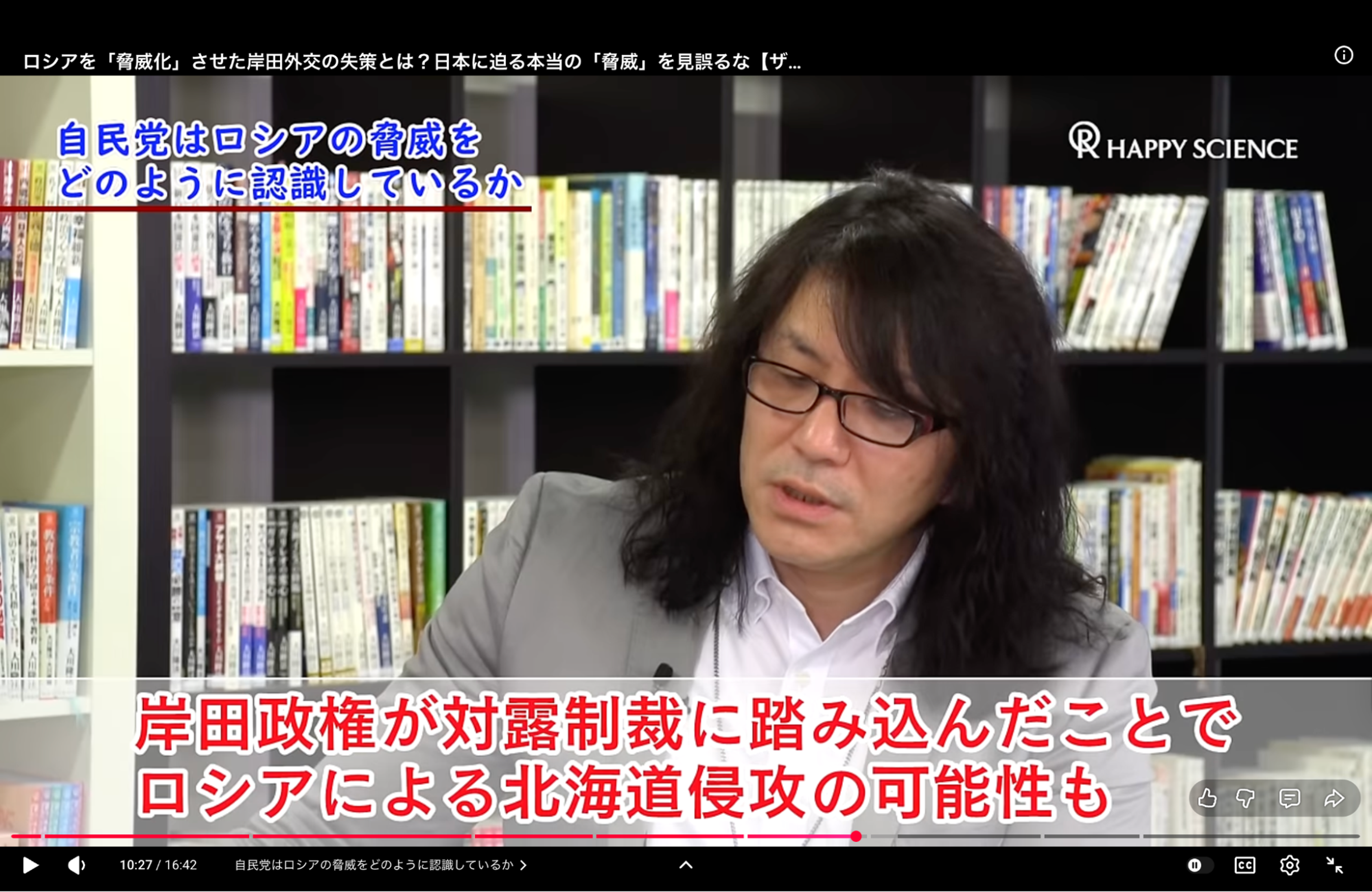 A man with glasses and long hair speaks in front of a bookshelf, with subtitles in Japanese text.
