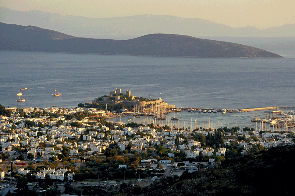 A view of a town beside the sea, taken from a hill. Many small white buildings, and a castle on a small promontory jutting out into the sea beside a harbour. Beyond a sea inlet are two ranges of hills.