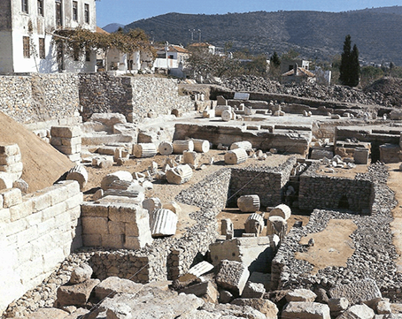 A view of the site of the Mausoleum in its present state, with stone walls from the monument’s foundations, and numerous drums from its columns. The background shows small houses and hills.
